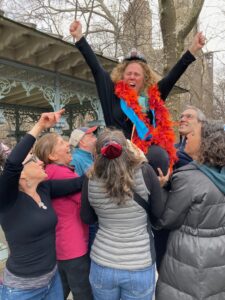 Devora is smiling while being lifted up in a chair by her family and friends in Central Park.